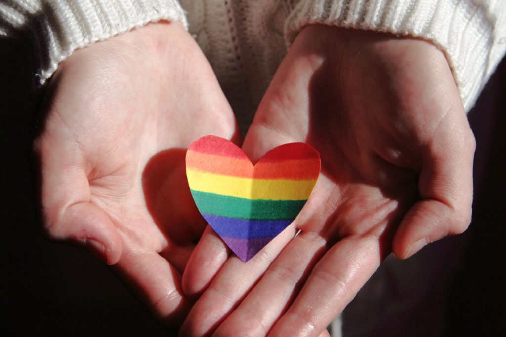 Two hands are cupped together holding a cut out of a rainbow heart