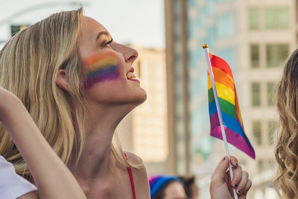 Image of a blond haired woman with rainbow face paint on her cheek waving a small rainbow flag