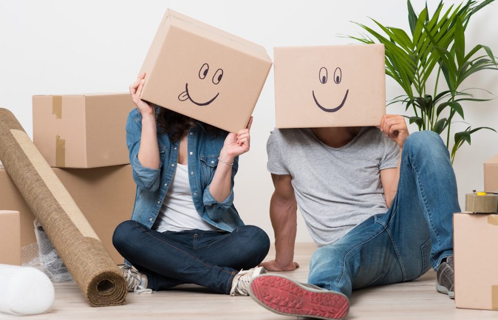 A pair of young people sit on the floor surrounded by boxes. They have boxes on their heads on to which they have drawn smiley faces.