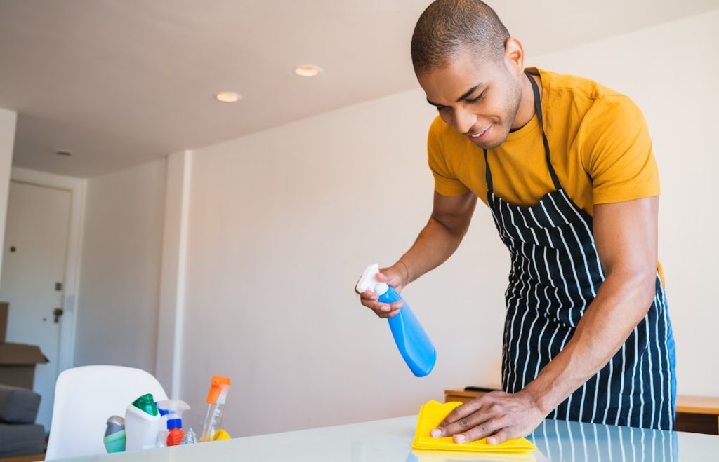A young person wearing an apron is cleaning a table with a cloth in one hand and a bottle of spray in the other.