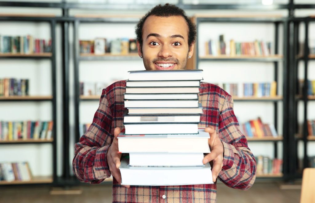 Stoke Student Living Blog post. Student Guide: De-clutter and make some money! 

A person stands smiling in front of some book shelves holding a large pile of books.