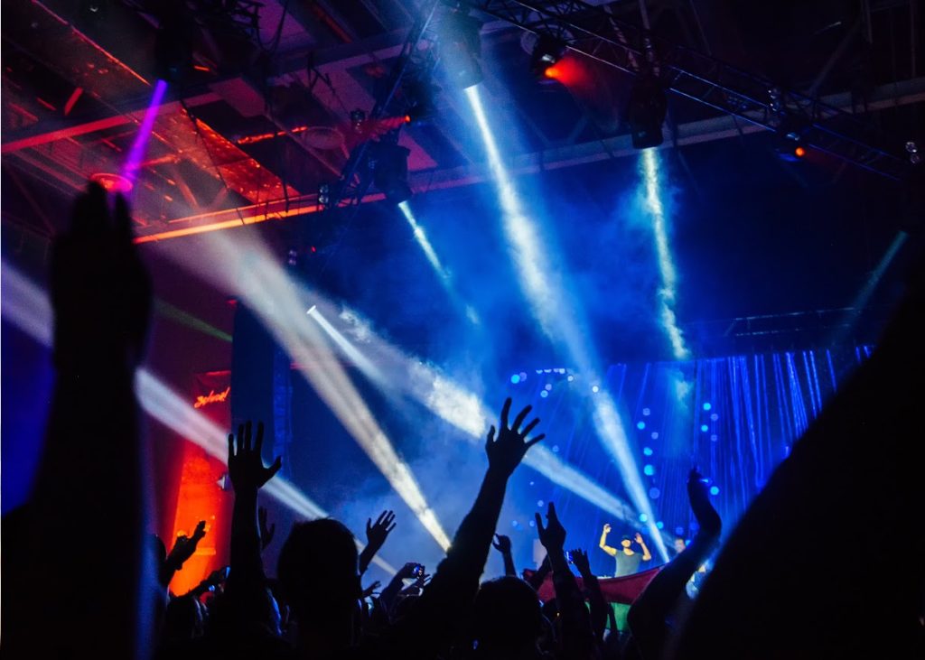 Hands raised high in the air in a 'rave' dance / nightclub setting with laser lights shining over the crowd.