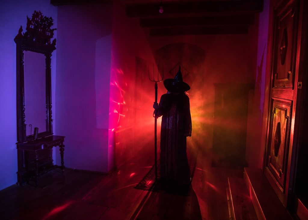 Spooky image of a silhouette of witch, there are coloured lights shining around the walls of the house she is standing in