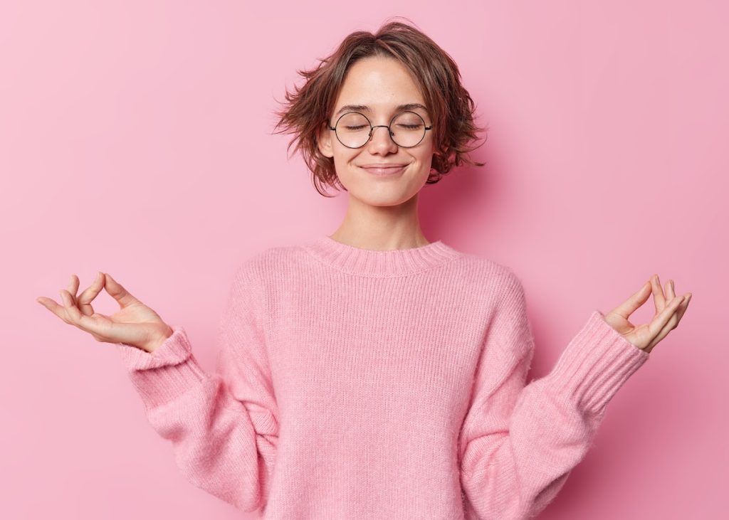 A young woman in a pink jumper stands against a pink background, with eyes closed and hands held in a meditation pose.