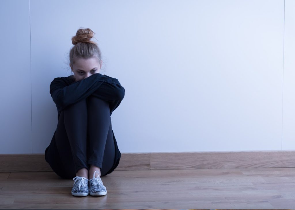 Worried young woman is sat on the floor against a wall with her knees drawn up and her head on her arms.