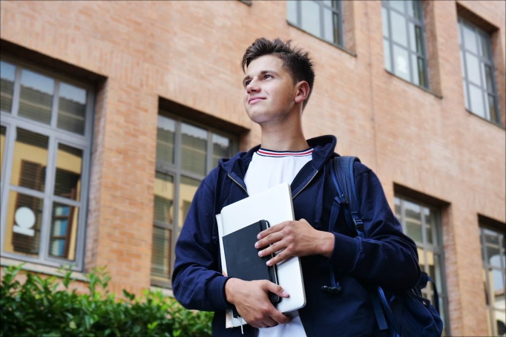 A young student holding some books and papers is seen looking into the distance in an outdoor setting.